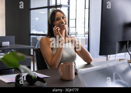 Donna che sorride e parla sullo smartphone alla scrivania in un ufficio open space con tazza da caffè e monitor. Professionale, spazio di lavoro, moderno Foto Stock