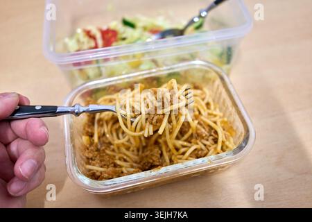Una persona mangia spaghetti bolognese da un contenitore di vetro con un'insalata di verdure fresca in una scatola di plastica dietro. Foto Stock