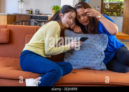Madre e figlia asiatiche sedute sul divano arancione a casa con lo smartphone appoggiato sul cuscino grigio. Accogliente, familiare, moderno, rilassante Foto Stock
