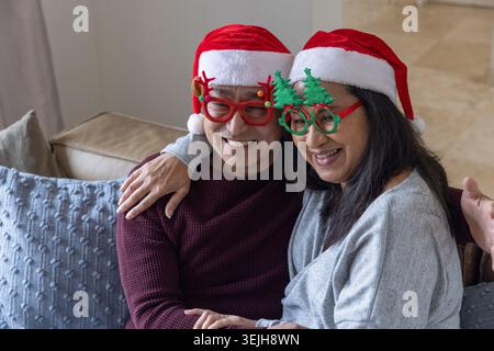 Diverse coppie di anziani sorridono indossando cappelli di Babbo Natale e occhiali natalizi sul divano nel soggiorno. Festa, festa, compagna Foto Stock