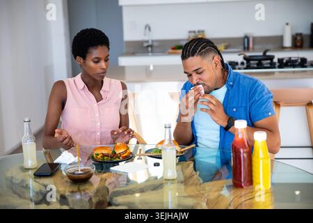 Coppie diverse che mangiano hamburger e insalata al tavolo da pranzo in vetro nella cucina moderna con condimenti. Contemporaneo, intimo, informale Foto Stock