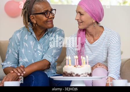 Diverse amiche che festeggiano il compleanno soffiando candele accese sulla torta sul divano del soggiorno. Festeggiamenti, compagnia, caldo Foto Stock