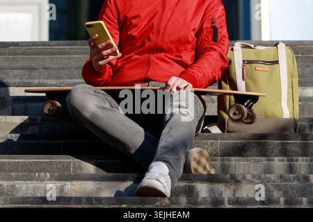 Uomo longboarder che usa il suo smartphone, riposa sui gradini, seduto con longboard, skateboard all'aperto Foto Stock