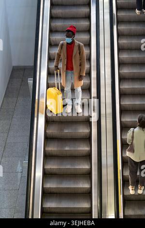 Uomo nero sta su scala mobile in aeroporto indossare maschera per proteggere dal contatto con il virus Nipah Foto Stock