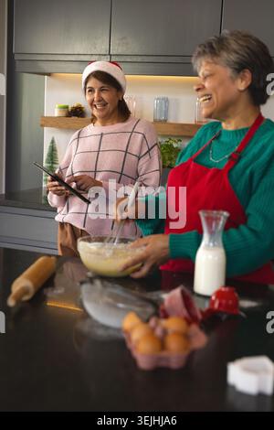 Amiche che preparano biscotti per le vacanze su un'isola di pietra in una cucina moderna utilizzando tablet e frusta. Festa, collaborazione, elega Foto Stock
