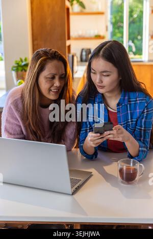 Madre e figlia asiatiche condividono lo smartphone mentre lavorano con un computer portatile al tavolo della cucina. Famiglia, legame, comunicazione, moderno, c Foto Stock