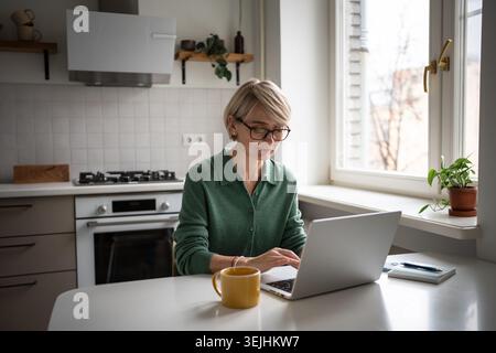Donna d'affari di mezza età, molto impegnata, che scrive su un notebook e lavora da casa al mattino. Foto Stock
