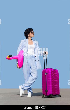 Ritratto estivo donna di mezza età in abiti casual, occhiali da sole con valigia e anello gonfiabile a forma di fenicottero ag Foto Stock