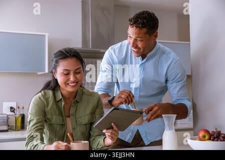 Coppia diversificata in piedi a casa cucina isola guardando tablet digitale e tazze mescolanti. Moderno, accogliente, vivace, collaborativo, stile di vita, famiglia, c Foto Stock