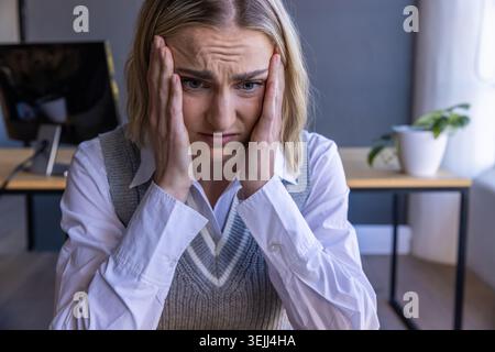 Donna che indossa un gilet grigio su una camicia bianca che tiene le aste alla scrivania dell'ufficio, accanto al monitor e alla pianta. Stress, concentrazione, spazio di lavoro, Foto Stock