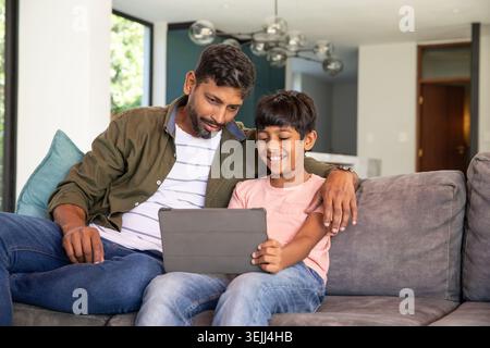 Padre e figlio indiani che indossano abiti casual seduti su un divano grigio usando un tablet nel soggiorno. Famiglia, relax, tecnologia, modernhome, legame, le Foto Stock