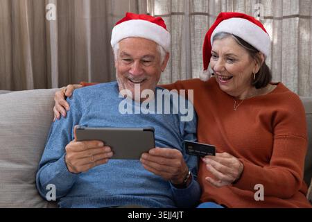 Una coppia anziana seduto sul divano nel soggiorno indossando cappelli di Babbo Natale con tablet e carta di credito. Anziani, relax, tecnologia, festa Foto Stock