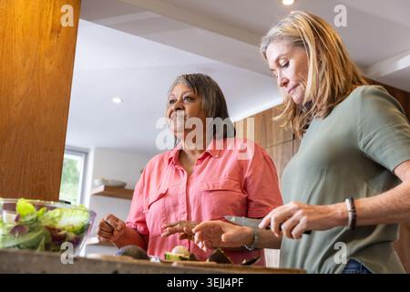 Diverse amiche tagliano metà di avocado sul tagliere al banco della cucina con ciotola per insalate. Cucinare, collaborare, guarire Foto Stock