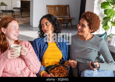 Diversi amici sono seduti sul divano nel soggiorno, tenendo in mano una tazza, una ciotola di pretzel e un telecomando. Accogliente, conviv Foto Stock