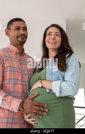 Coppie diverse che mettono le mani sul baule del bambino e si mettono in piedi in una casa vicino alla ringhiera delle scale. Gravidanza, famiglia, mothe Foto Stock