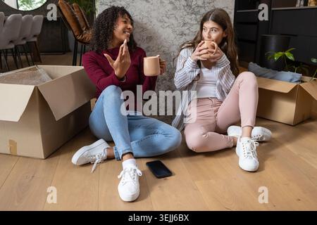 Vari amici chiacchierano e disimballano le scatole a casa vicino al loro smartphone tenendo in mano tazze in ceramica. Intimità, amico Foto Stock