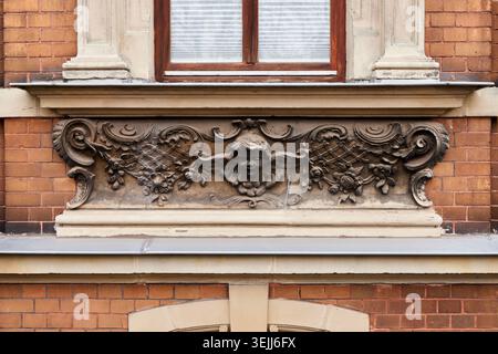 Dettaglio architettonico di un vecchio edificio in mattoni caratterizzato da un rilievo in pietra ornato con una faccia d'angelo cherubino e motivi floreali sotto una finestra. Foto Stock