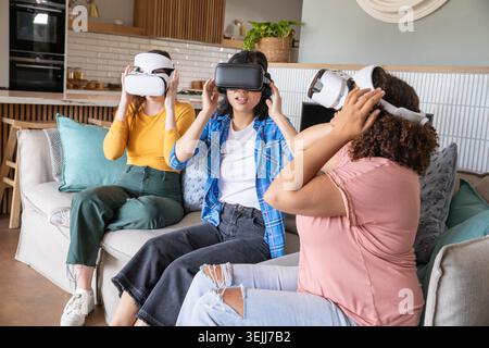Diverse amiche stanno provando i visori VR mentre sono sedute su un divano con cuscini accanto alla cucina. Innovativo, rilassato, Foto Stock