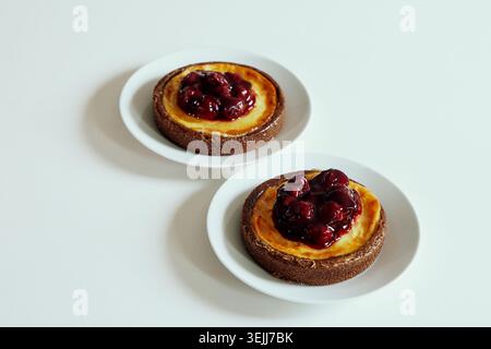 Due crostate di cheesecake alla ciliegia condite con marmellata di frutti rossi lucida, servite su piatti di ceramica bianca separati su uno sfondo pulito. Foto Stock