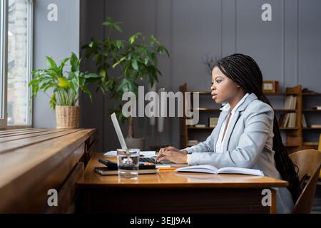 Donna nera d'affari concentrata in tuta da custode di libri remoti che prepara report finanziari su un notebook Foto Stock