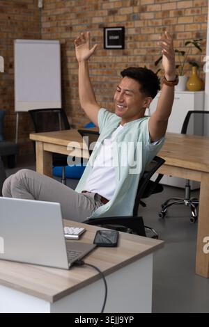 Uomo asiatico che si allunga le braccia sopra la testa seduto alla scrivania in ufficio con la lavagna a fogli mobili dello smartphone del computer portatile. Motivato, energico, moderno, W Foto Stock
