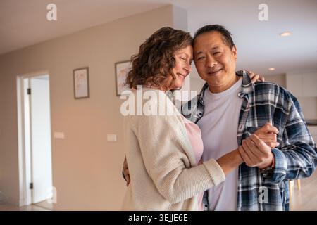 Diverse coppie senior che ballano lentamente in un'area soggiorno open space con stampe incorniciate e cucina isolana. Romanticismo, intimità, affetto Foto Stock