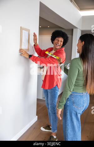 Diverse coppie che appendono una cornice in legno sulla parete di una casa moderna utilizzando una livella a bolla gialla. Collaborazione, interni, moderno, d Foto Stock