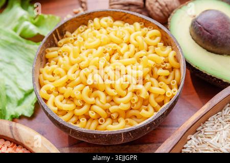 Pasta cruda macaroni in una ciotola di legno su una superficie rustica con cibo diverso. Foto Stock