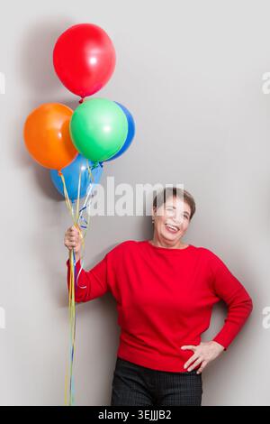 Signora anziana felice che tiene regali e palloncini multicolori che guardano la macchina fotografica e sorridono in posa intorno a un muro. Natale. B Foto Stock