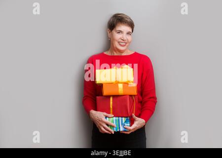 Una donna anziana felice che tiene regali guardando la macchina fotografica e sorridendo che posa su un muro. Natale. Compleanno Foto Stock