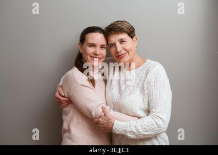 Felice signora madre anziana e figlia adulta che abbracciano guardando la macchina fotografica su uno sfondo grigio Foto Stock