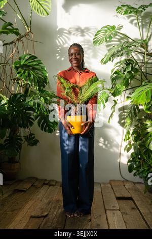 Una donna afroamericana sorridente tiene Cycas in vaso, circondata da lussureggianti giardini tropicali. Foto Stock