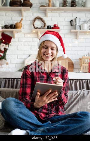 Giovane donna bionda nel cappello di babbo natale che lavora su un tablet seduto sul divano Foto Stock