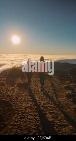 Due amici camminano su un sentiero di montagna sopra le nuvole, tenendosi per mano durante il tramonto Foto Stock