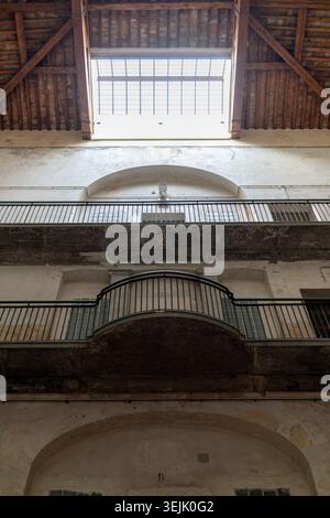 Una vista interna di un vecchio edificio, che presenta diversi balconi con ringhiere e una struttura rustica a soffitto in legno che conduce ad un luminoso lucernario Foto Stock
