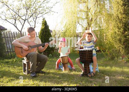 Famiglia felice papà e due bambini che si divertono insieme con strumenti musicali all'aperto. Papà che suona la chitarra e i bambini che suonano etnico Foto Stock