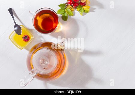Tè alla rosa con teiera, tazza, miele, ramo d'anca di rosa d'autunno in giornata di sole con ombre su tavola di legno bianco vista dall'alto, spazio copia. Foto Stock