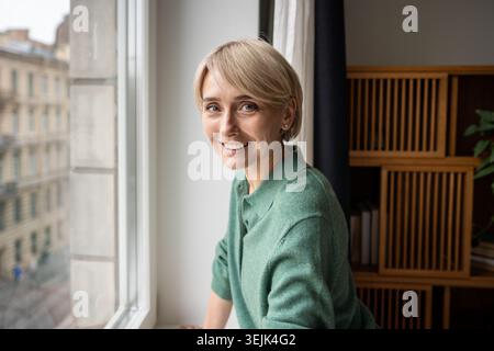 Donna allegra con un'espressione positiva che posa vicino alla finestra e guarda la fotocamera con un sorriso felice. Foto Stock
