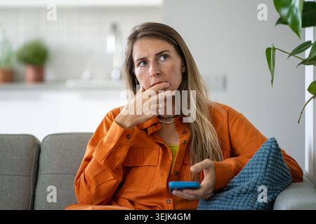 Donna di mezza età che pensa profondamente fornendo analisi approfondite tenendo il telefono seduto sul divano di casa Foto Stock
