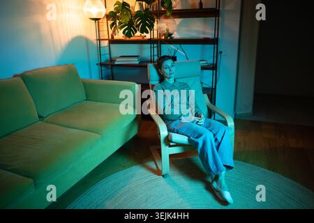Una donna anziana siede sulla sedia di casa con luce rgb guardando con apatia, tristezza e stanchezza mentale. Foto Stock