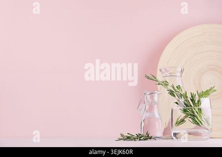 Ramoscelli di rosmarino verde in vaso di vetro trasparente e piatto di bambù beige su morbido sfondo rosa pastello. Fresca stagione con dorso delicato Foto Stock