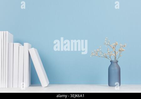 Elegante arredamento per la casa - libri bianchi e piccolo vaso in vetro con fiori secchi su un morbido tavolo in legno bianco chiaro e sfondo blu. Foto Stock