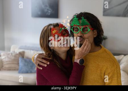 Coppia che indossa occhiali natalizi, si accoccolano sul divano in soggiorno, tengono i volti, smartwatch visibile. Accogliente, caldo, romantico, coppia, h Foto Stock