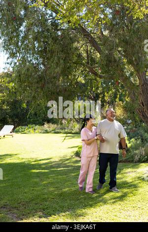 Infermiera e uomo diversi che camminavano sotto l'albero ombra in giardino, scrub aiutavano vicino alla poltrona bianca Foto Stock