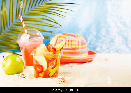Macedonia di frutta fresca in scatola trasparente da asporto con cocktail freddo, mela verde sulla spiaggia tropicale estiva con palme, sabbia, soleggiata Foto Stock