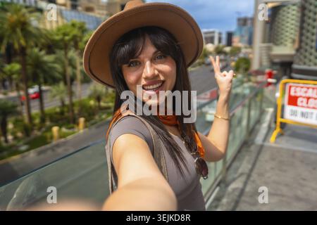 Donna con cappello e sciarpa che si gode la sua vacanza a Las vegas, nevada, sorridendo alla macchina fotografica e facendo un gesto di pace mentre cattura un memorabile Foto Stock