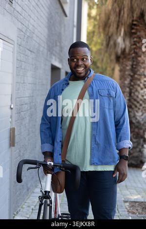 Uomo afro-americano che indossa una camicia in denim che tiene in mano la bicicletta da strada e la borsa a tracolla sul vialetto pavimentato. Urbano, marciapiede, ciclismo, casu Foto Stock