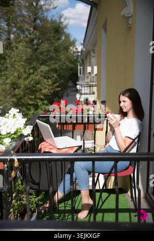 Giovane donna che lavora con un portatile e beve una tazza di tè sul balcone del suo appartamento in una giornata estiva. Foto Stock