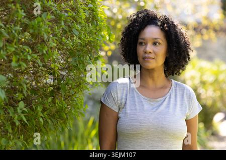 Donna afroamericana in piedi accanto alla fitta siepe verde nel giardino illuminato dal sole e indossa una T-shirt grigia Foto Stock