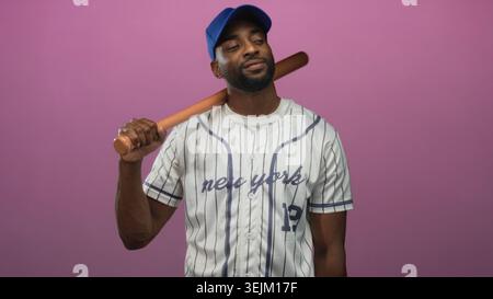 Uomo che tiene una mazza da baseball sulla spalla in uno studio con indosso una maglia di tessuto e un berretto blu, il numero 12 visibile, posa rilassata e un leggero sorriso; tè di fiducia Foto Stock